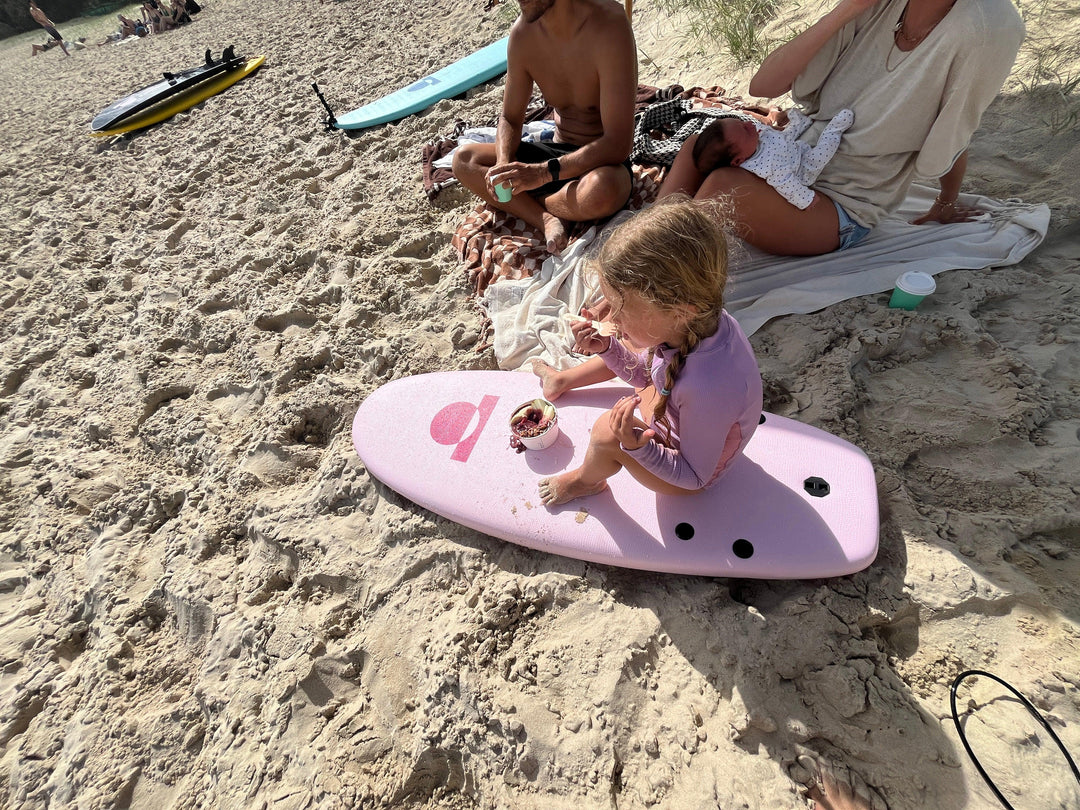 Child sitting on a pink surfboard on a sandy beach with people in the background.